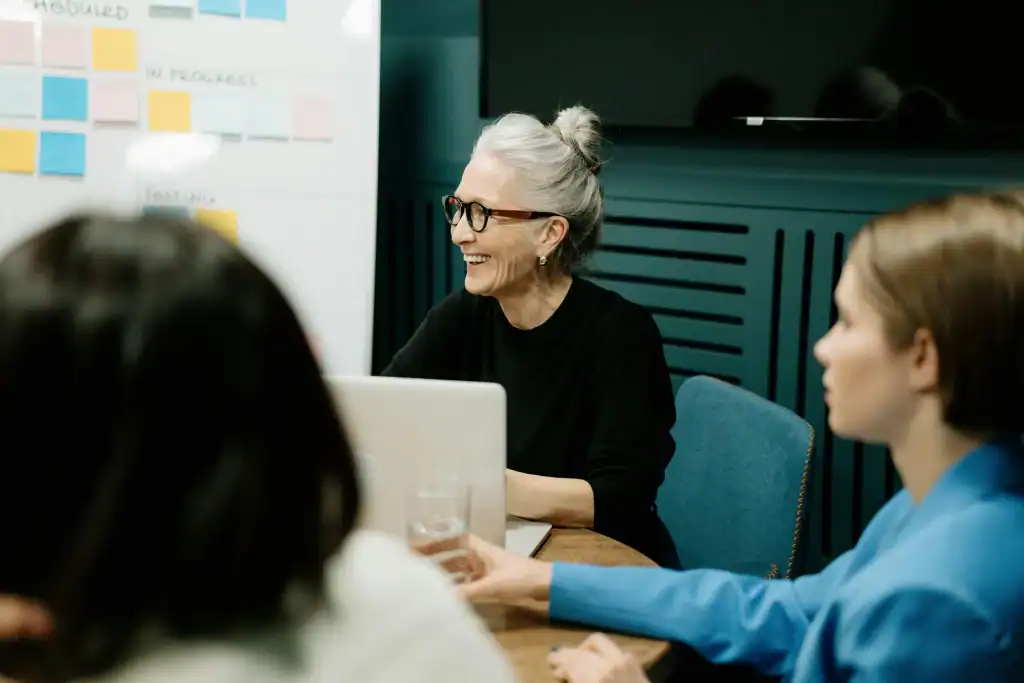 Team members in a meeting, with a smiling woman at a laptop and sticky notes on a whiteboard in the background.