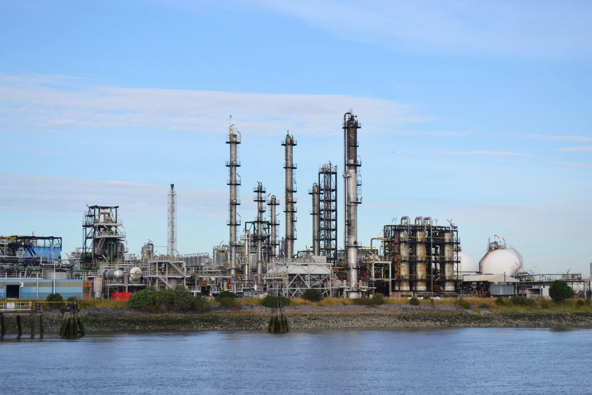 Refinery or chemical plant with distillation columns and pressure vessels, viewed from across a water channel under clear blue skies.