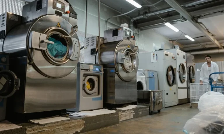 A row of large stainless steel industrial washer-extractors on the floor of a commercial laundry plant, with dryers further down the line