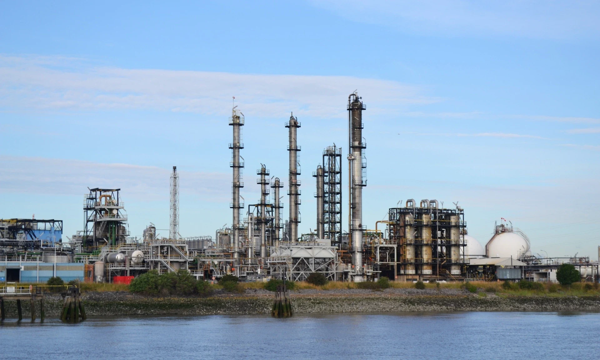 Refinery or chemical plant with distillation columns and pressure vessels, viewed from across a water channel under clear blue skies.